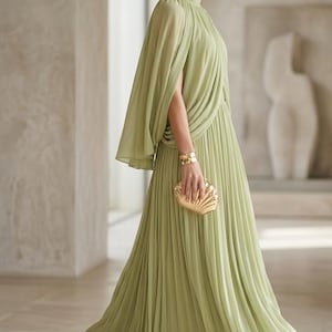 Puede incluir: Un vestido largo de color verde salvia con cuello alto, falda plisada y una capa superpuesta integrada. La prenda se complementa con pendientes circulares dorados, un brazalete met&aacute;lico ancho y un bolso de mano dorado con forma de concha.