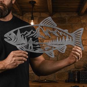 May include: A flat, dark gray metal wall art piece shaped like a fish. The cutout design inside the fish silhouette features a detailed landscape with a person fishing in a lake, surrounded by a dense forest of pine trees and jagged mountains.