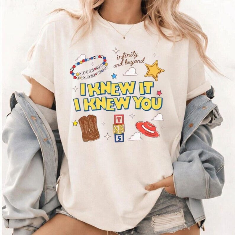 K&ouml;nnte beinhalten: Ein cremefarbenes T-Shirt mit einem Grafikdruck, der den Text "I KNEW IT I KNEW YOU" und "infinity and beyond" zeigt. Das Design enth&auml;lt Illustrationen eines Perlenarmbands, eines gelben Sterns, brauner Cowboystiefel, Spielw&uuml;rfel und eines roten Hutes.