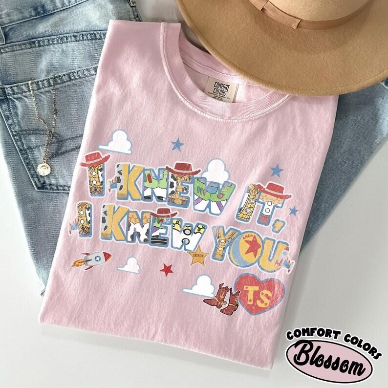 May include: A light pink t-shirt featuring the text I KNEW IT, I KNEW YOU in a block font filled with patterns inspired by cowboy and space ranger characters. The design includes small clouds, stars, a rocket, a heart with the letters TS, and boots.