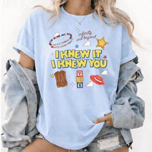 K&ouml;nnte beinhalten: Ein hellblaues T-Shirt mit dem gelben Schriftzug I KNEW IT I KNEW YOU sowie Grafiken eines Perlenarmbands, eines goldenen Sterns, brauner Cowboystiefel, eines Stapels Buchstabenw&uuml;rfel und eines roten Hutes, mit dem Text infinity and beyond.