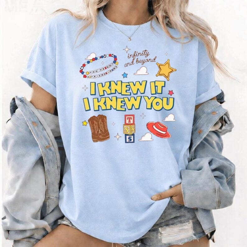 K&ouml;nnte beinhalten: Ein hellblaues T-Shirt mit dem gelben Schriftzug I KNEW IT I KNEW YOU sowie Grafiken eines Perlenarmbands, eines goldenen Sterns, brauner Cowboystiefel, eines Stapels Buchstabenw&uuml;rfel und eines roten Hutes, mit dem Text infinity and beyond.
