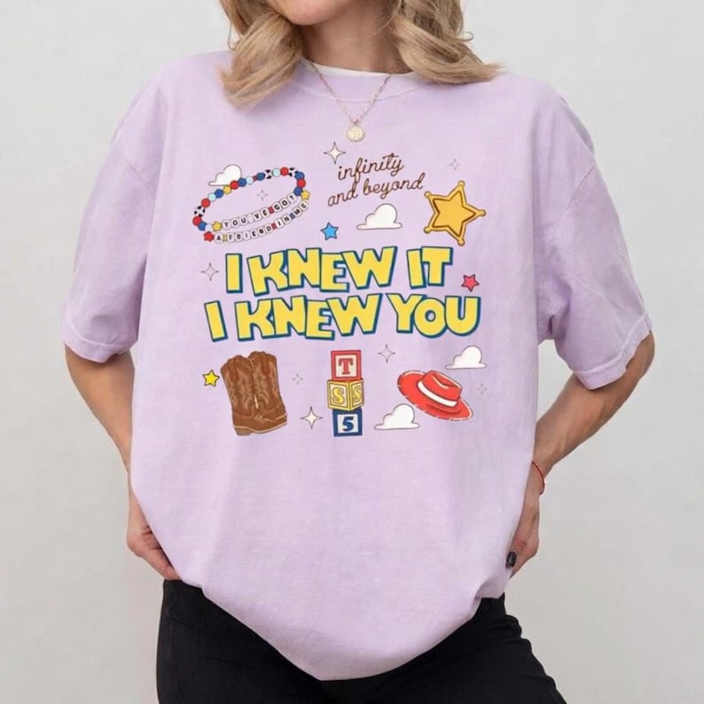 K&ouml;nnte beinhalten: Ein hellviolettes T-Shirt mit einem Grafikdruck mit dem Text "I KNEW IT I KNEW YOU" und "infinity and beyond". Das Design enth&auml;lt Illustrationen eines Perlenarmbands, eines gelben Sterns, brauner Cowboystiefel, Buchstabenw&uuml;rfel und eines roten Hutes.