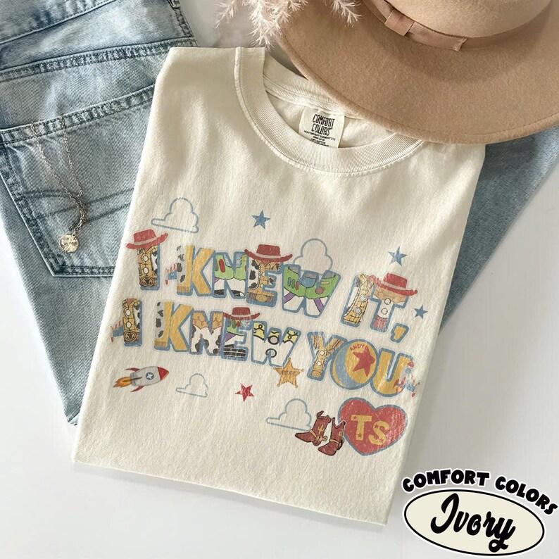 May include: An ivory t-shirt featuring the text I KNEW IT, I KNEW YOU in a multi-colored font with patterns inspired by cowboy and space ranger characters. The design includes small stars, clouds, a rocket, cowboy boots, and a heart with the letters TS.