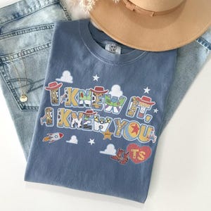 May include: A blue t-shirt featuring the text I KNEW IT, I KNEW YOU in a multi-colored graphic print with cowboy hat, space ranger, and sheriff badge motifs, along with a red heart containing the letters TS, white clouds, and stars.