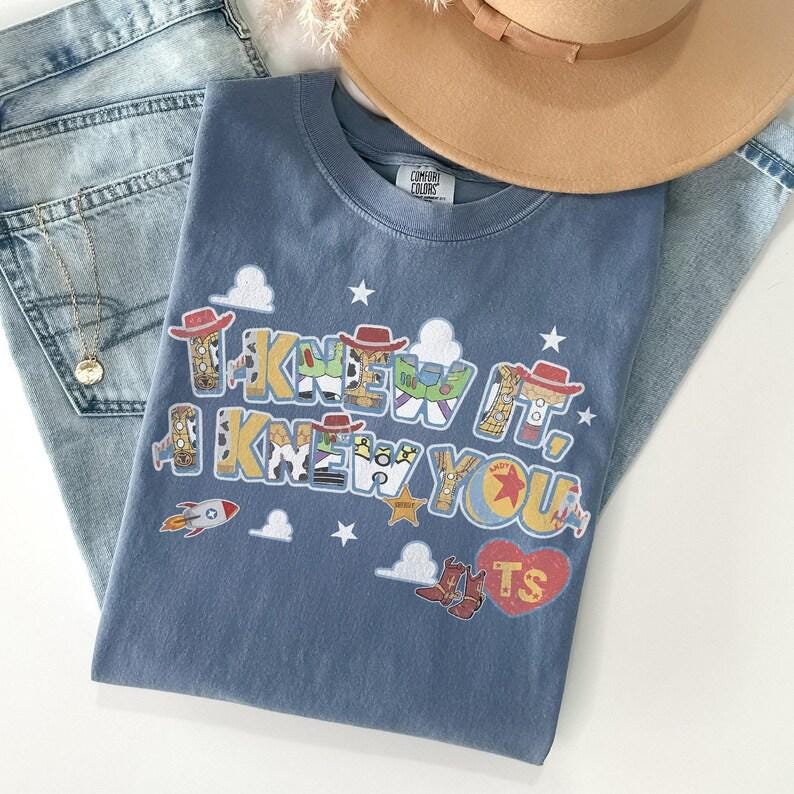 May include: A blue t-shirt featuring the text I KNEW IT, I KNEW YOU in a multi-colored graphic print with cowboy hat, space ranger, and sheriff badge motifs, along with a red heart containing the letters TS, white clouds, and stars.