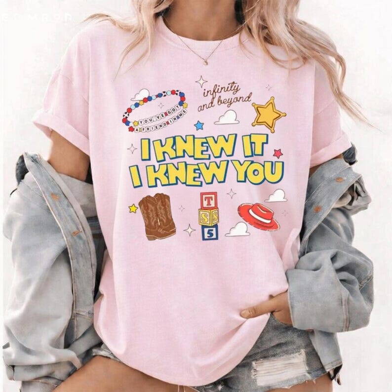 K&ouml;nnte beinhalten: Ein hellrosa T-Shirt mit dem Text I KNEW IT I KNEW YOU in Gelb mit blauen Umrandungen. Die Grafiken zeigen ein Perlenarmband, einen goldenen Stern, braune Cowboystiefel, drei gestapelte Buchstabenw&uuml;rfel und einen roten Cowboyhut mit Wolken.
