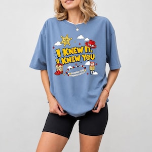 May include: A blue short-sleeve t-shirt featuring a graphic print with the text "I KNEW IT, KNEW YOU" in yellow. The design includes a yellow sheriff star, a red cowboy hat, red cowboy boots, a beaded bracelet, and alphabet blocks with the letter T and 5.