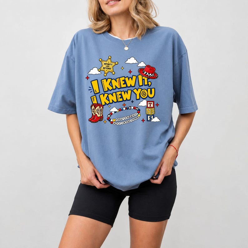 May include: A blue short-sleeve t-shirt featuring a graphic print with the text "I KNEW IT, KNEW YOU" in yellow. The design includes a yellow sheriff star, a red cowboy hat, red cowboy boots, a beaded bracelet, and alphabet blocks with the letter T and 5.