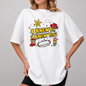 May include: A white t-shirt featuring a graphic print with the text I KNEW IT, I KNEW YOU. The design includes a yellow sheriff badge, a red cowboy hat, red cowboy boots, a colorful beaded bracelet with text, and three stacked alphabet blocks.