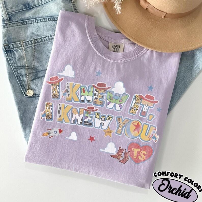 May include: A light purple t-shirt featuring the text I KNEW IT, I KNEW YOU in a multi-colored font with character-inspired patterns. The design includes small cloud, star, rocket, and heart graphics, with a red heart containing the letters TS.
