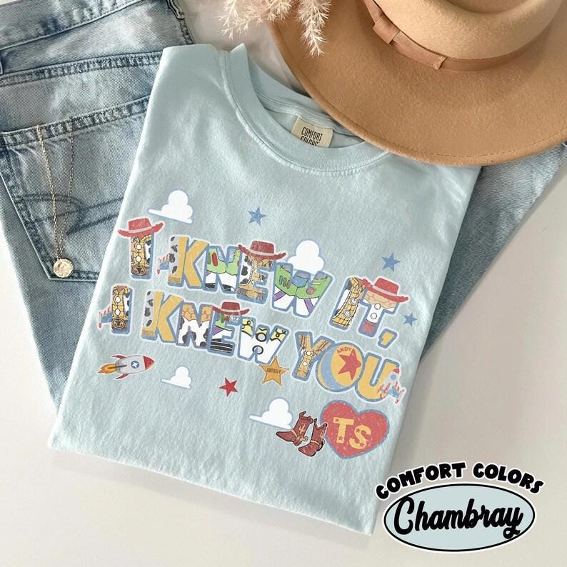 May include: A light blue t-shirt featuring the text I KNEW IT, I KNEW YOU in a multi-colored font filled with patterns of cowboy hats, space ranger suits, and cow print. The design includes small rocket, star, cloud, and heart graphics.