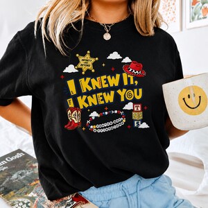 May include: A black t-shirt featuring a graphic print with the yellow text I KNEW IT, I KNEW YOU. The design includes a gold sheriff badge, a red cowboy hat, red cowboy boots, a beaded bracelet with the text YOU'VE GOT A FRIEND IN ME, and blocks.