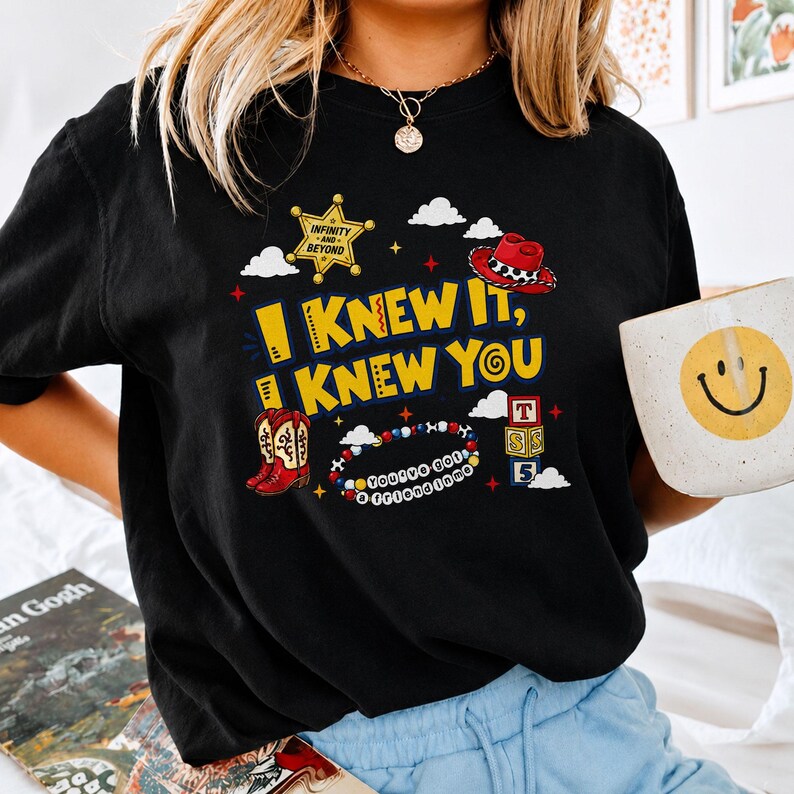 May include: A black t-shirt featuring a graphic print with the yellow text I KNEW IT, I KNEW YOU. The design includes a gold sheriff badge, a red cowboy hat, red cowboy boots, a beaded bracelet with the text YOU'VE GOT A FRIEND IN ME, and blocks.