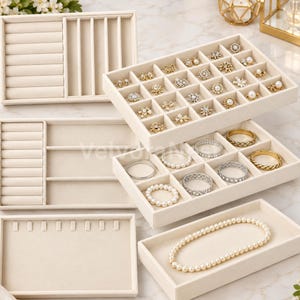 May include: A set of beige velvet jewelry organizer trays featuring various compartments for rings, earrings, and necklaces. The trays include slotted sections for rings and open square grids for small accessories, with a pearl necklace displayed in one tray.