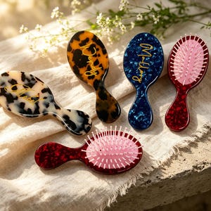 May include: Five oval hairbrushes with handles, featuring various tortoiseshell patterns in cream, brown, blue, and red. Each brush has a glossy finish and gold cursive text on the back. Two brushes show pink bristles, while others are face down.