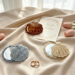 Könnte beinhalten: Drei muschelförmige Taschenspiegel in Braun, Hellblau und Creme, jeweils mit goldfarbenen Konturen. Die Spiegel sind geöffnet und zeigen eine reflektierende Fläche. Ebenfalls enthalten sind zwei goldfarbene Ringe, eine Karte mit der Aufschrift B&M Wedding und ein Lippenstift.