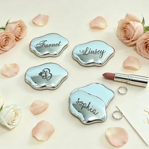 Pode incluir: Quatro espelhos de bolso prateados de formato irregular, semelhante a uma nuvem, com gravações personalizadas lendo Farnet, Linsey, B e Sophia. Um batom, dois anéis prateados e um cartão com o texto S&Y Wedding entre pétalas de rosa pálidas.