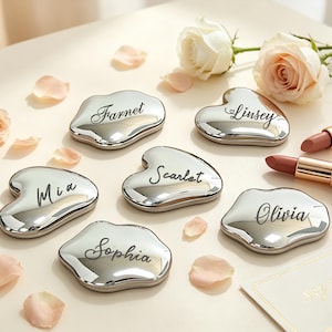 May include: Six silver-toned, irregular heart-shaped compact mirrors, each engraved with a unique name: Farnet, Linsey, Mia, Scarlet, Olivia, and Sophia. The mirrors are arranged alongside two lipsticks and several pale pink rose petals.