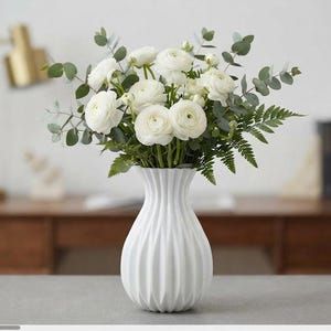 May include: A white, vertically ribbed vase holds a bouquet of white ranunculus flowers, green eucalyptus sprigs, and fern leaves. The vase has a narrow neck that flares out into a rounded, textured body with deep, vertical grooves throughout.