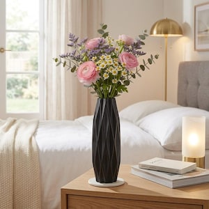 May include: A tall, matte black vase with a geometric, faceted surface texture. The vase holds a vibrant floral arrangement featuring pink ranunculus, small white daisies, purple statice, and green eucalyptus stems, creating a contrast.