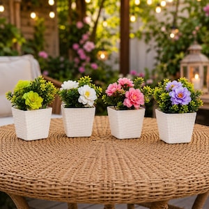May include: Four small square white pots with textured surfaces, each containing a different artificial flower arrangement. The flowers are lime green, white, pink, and purple, all surrounded by small green leaves, arranged in a row on a table.