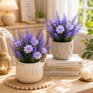 May include: Two artificial lavender arrangements in white ceramic pots. Each pot contains purple lavender sprigs and light purple flowers. One pot is ribbed, the other has a geometric pattern. They sit on a woven mat and stacked books.