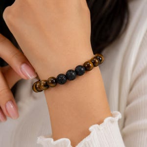Puede incluir: Una pulsera de cuentas que presenta una alternancia de cuentas de piedra ojo de tigre de color marrón dorado liso y cuentas de piedra volcánica negra porosa y texturizada. Las cuentas esféricas forman un accesorio de muñeca.