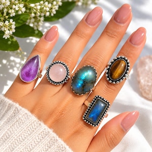 Puede incluir: Cinco anillos de plata con diversas piedras preciosas: una amatista morada en forma de pera, un cuarzo rosa pálido redondo, una gran labradorita ovalada iridiscente, un ojo de tigre ovalado marrón y una labradorita azul rectangular, todos con engastes de cuentas.