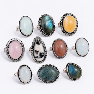 Puede incluir: Una colección de nueve anillos con piedras preciosas cabujón de forma ovalada y de lágrima, engastadas en monturas de metal plateado decorativas. Las piedras presentan varios colores como azul pálido, azul oscuro iridiscente, amarillo, rosa, blanco y patrones moteados en blanco y negro.