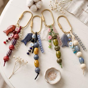 May include: Four gold-toned metal oval keychains, each featuring a fish-shaped charm and a vertical arrangement of multi-coloured beads. The beads include various textures, patterns, and sizes in red, blue, green, and cream, accented with small crystal-like strands.