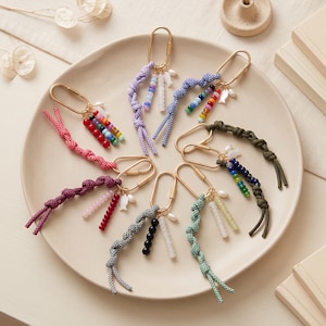 May include: A collection of seven oval gold-tone metal carabiner keychains, each featuring a knotted cord in various colours and patterns, accented with small colourful beads, a single white pearl, and a small white fish-shaped charm.