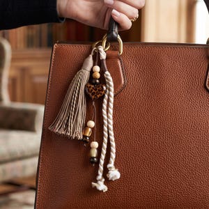 Könnte beinhalten: Eine braune Ledertasche mit einem dekorativen Anhänger am Griff. Der Anhänger besteht aus einer beigen Quaste, einem herzförmigen Anhänger mit Leopardenmuster und zwei gedrehten beigeweißen Kordeln mit Holzperlen.