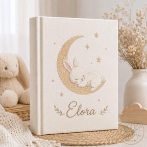 May include: A cream-colored fabric-covered book featuring gold-toned embroidery of a sleeping rabbit resting on a crescent moon, surrounded by small stars. The name Elora is embroidered in a cursive script below the moon with decorative leaf accents.