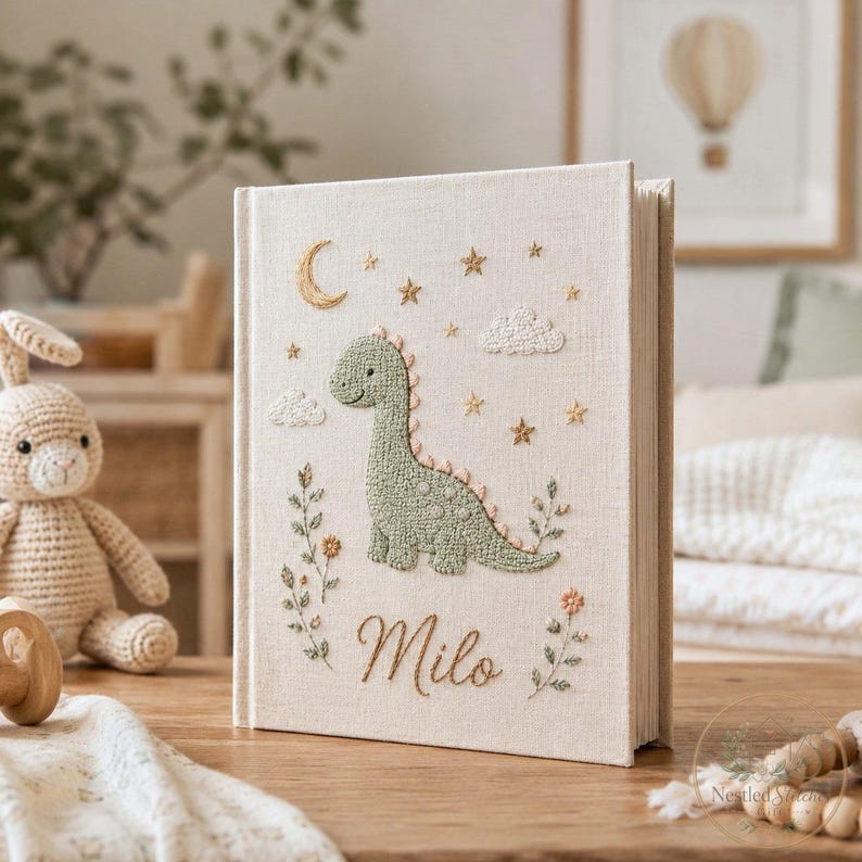 May include: A cream-colored fabric-covered book featuring an embroidered green dinosaur, a crescent moon, stars, and clouds. The name Milo is embroidered in cursive script at the bottom, flanked by two floral stems with small orange and green leaves.