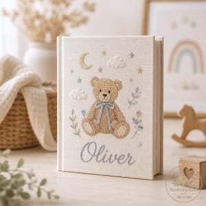 May include: A cream-colored fabric-covered book featuring an embroidered design of a brown teddy bear with a blue ribbon, surrounded by clouds, stars, and floral sprigs. The name Oliver is embroidered in blue script text at the bottom center.