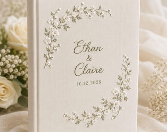 May include: A cream-colored fabric-covered book featuring embroidered white floral vines and green leaves. The center text reads Ethan & Claire with the date 10.12.2026 in a dark green script font. The cover has a hardbound spine and textured finish.