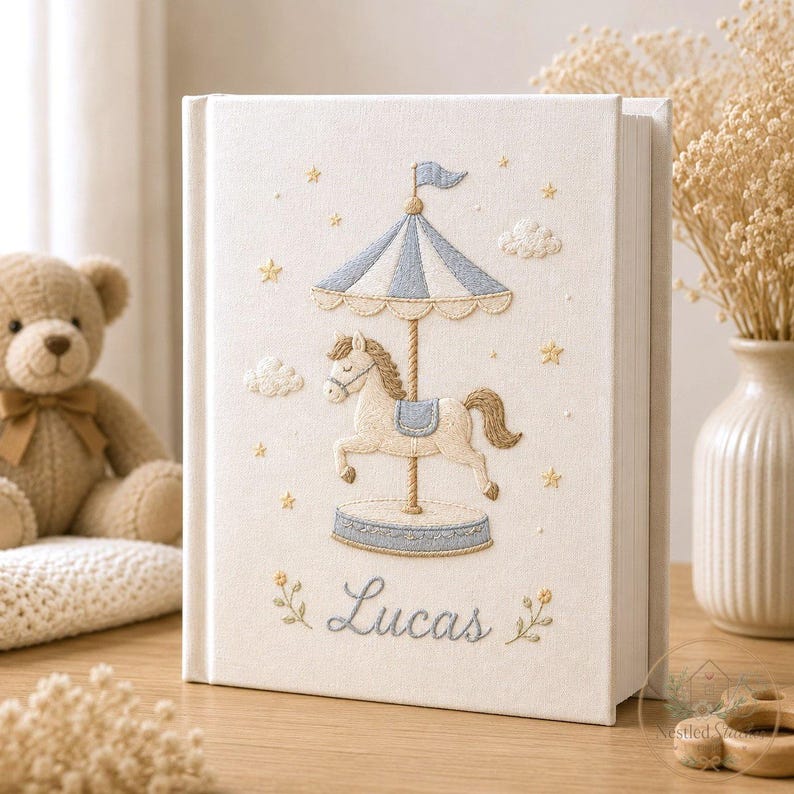 May include: A cream-colored fabric-covered book featuring an embroidered carousel horse under a blue and white striped canopy. The design includes small yellow stars, clouds, and the name Lucas stitched in blue cursive script at the bottom.