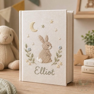 May include: A cream-colored fabric-covered book featuring embroidered details of a brown rabbit, a yellow crescent moon, and small stars. The cover includes green floral stems with blue and yellow flowers and the name Elliot stitched in green.