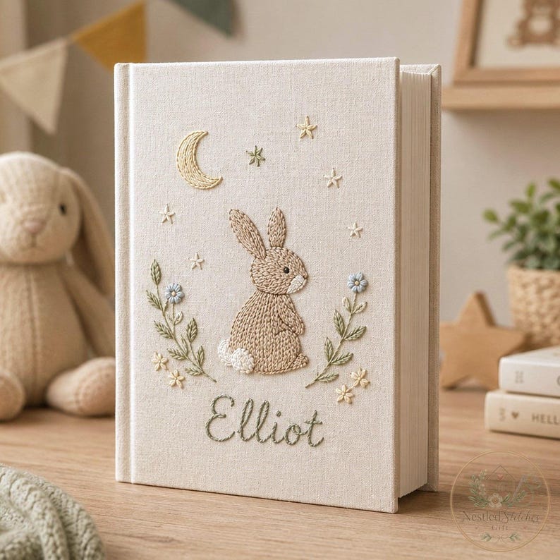 May include: A cream-colored fabric-covered book featuring embroidered details of a brown rabbit, a yellow crescent moon, and small stars. The cover includes green floral stems with blue and yellow flowers and the name Elliot stitched in green.