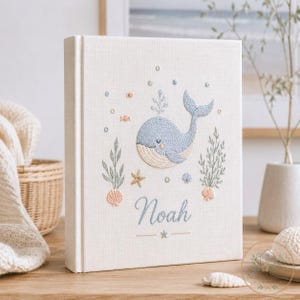 May include: A cream-colored fabric-covered book featuring an embroidered light blue whale, sea plants, seashells, and a starfish. The name Noah is embroidered in blue script below the whale, with small bubbles and a star accenting the design.