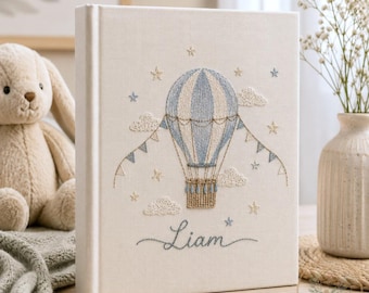 May include: A cream-colored fabric-covered photo album featuring an embroidered design of a blue and white hot air balloon, clouds, and stars. The name Liam is embroidered in cursive script below the balloon, accompanied by decorative bunting.