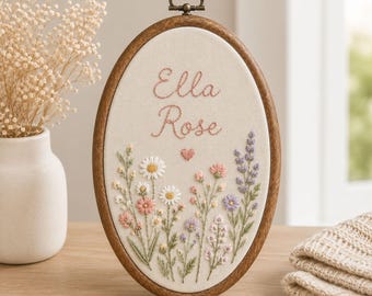 May include: An oval embroidery hoop featuring a dark wood frame and a cream-colored fabric center. The fabric displays the embroidered name Ella Rose in pink script, a small pink heart, and a colorful arrangement of various wildflowers and stems.