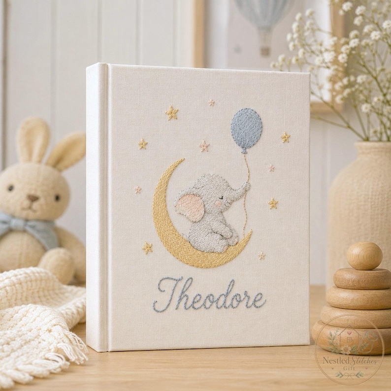 May include: A cream-colored fabric-covered photo album featuring an embroidered design of a gray elephant sitting on a yellow crescent moon, holding a blue balloon. The name Theodore is embroidered in blue script below the moon and stars.
