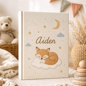 May include: A cream-colored fabric-covered book featuring embroidered details of a sleeping fox on a cloud, a crescent moon, stars, and clouds. The name Aiden is embroidered in brown script in the center of the cover above the fox.