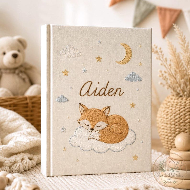 May include: A cream-colored fabric-covered book featuring embroidered details of a sleeping fox on a cloud, a crescent moon, stars, and clouds. The name Aiden is embroidered in brown script in the center of the cover above the fox.