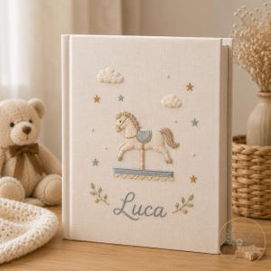 May include: A cream-colored fabric-covered photo album featuring embroidered details of a carousel horse, clouds, and stars. The name Luca is embroidered in blue script at the bottom, flanked by small green leafy sprigs and yellow accents.