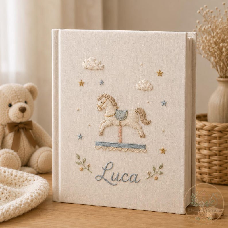 May include: A cream-colored fabric-covered photo album featuring embroidered details of a carousel horse, clouds, and stars. The name Luca is embroidered in blue script at the bottom, flanked by small green leafy sprigs and yellow accents.