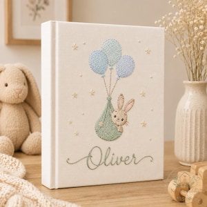 May include: A white fabric-covered memory book featuring an embroidered design of a light brown bunny inside a sage green sack held by three blue balloons. The name Oliver is embroidered in sage green cursive script below the illustration.