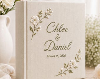 May include: A cream-colored fabric photo album featuring embroidered green floral sprigs and the text "Chloe & Daniel" followed by the date "March 15, 2026" in green thread. The cover has a textured linen appearance with a vertical spine detail.